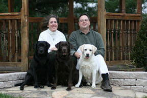 black, chocolate & yellow labs sitting with man & woman outside