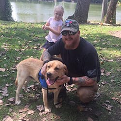 yellow Labrador service dog standing in grass with his handler wearing a baseball cap & glasses with a young girl, trees and a lake behind