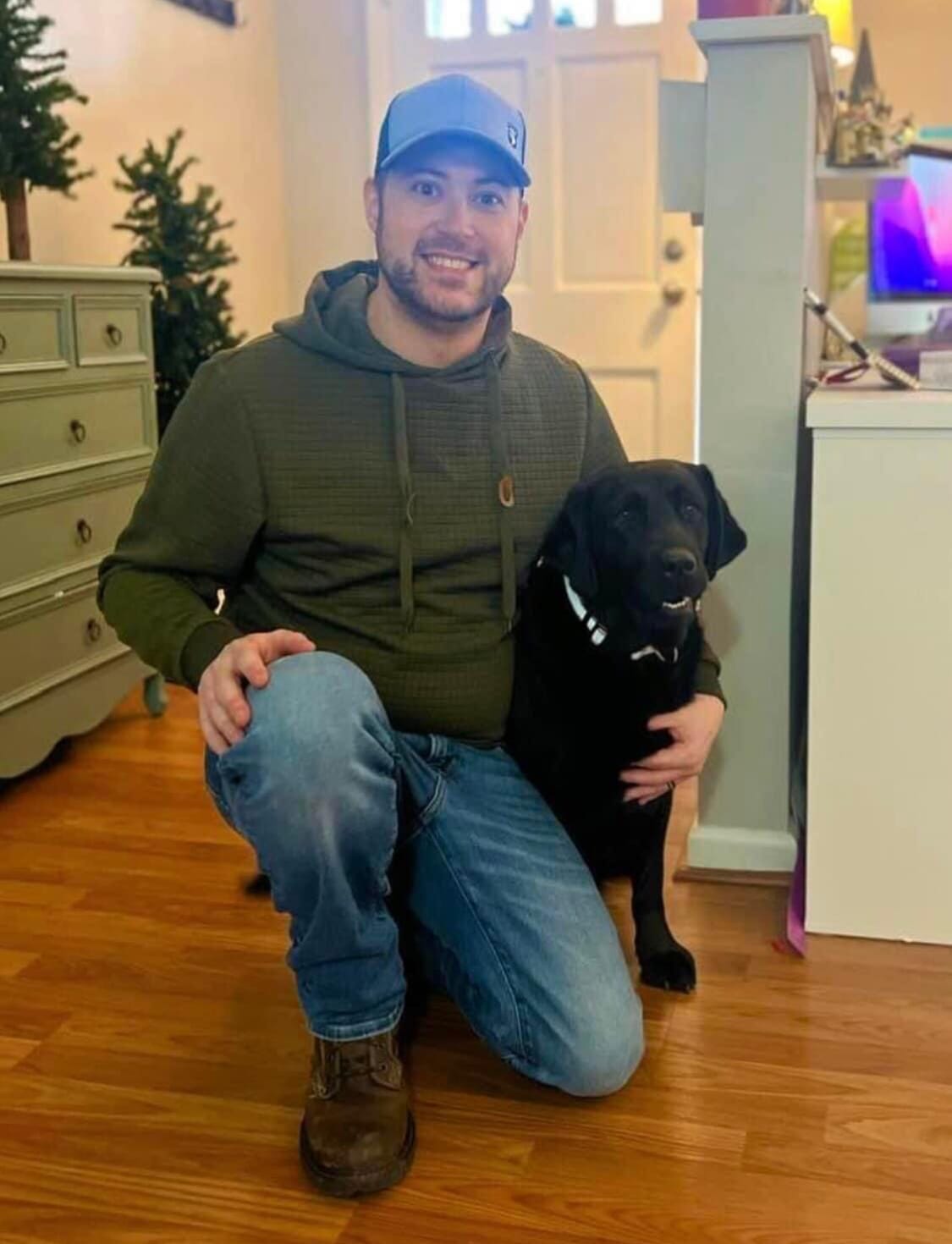 Man with his arm around his black Labrador Service dog man is wearhing jeans, a dark sweatshirt kneeling on a hardwood floor