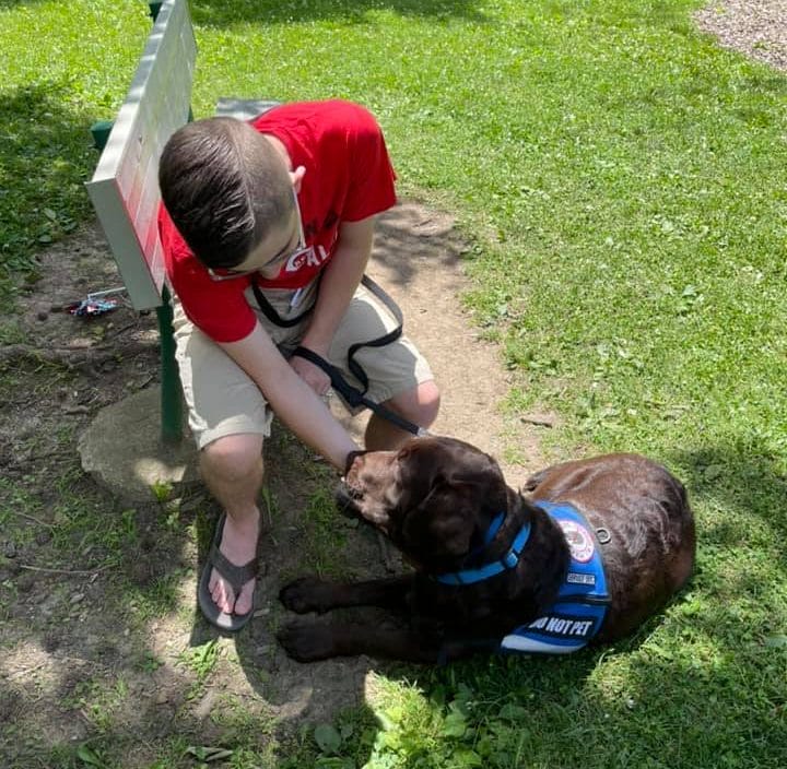 young man sitting on a park bench wearing a red shirt & khaki shorts with his chocolate Labrador service dog laying at his feet