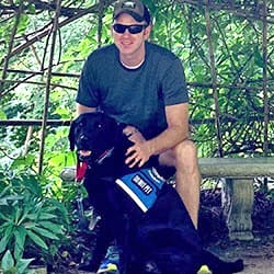 man wearing baseball hat in a dark blue t-shirt sitting on a bench with his black Labrador service dog sitting in front of him